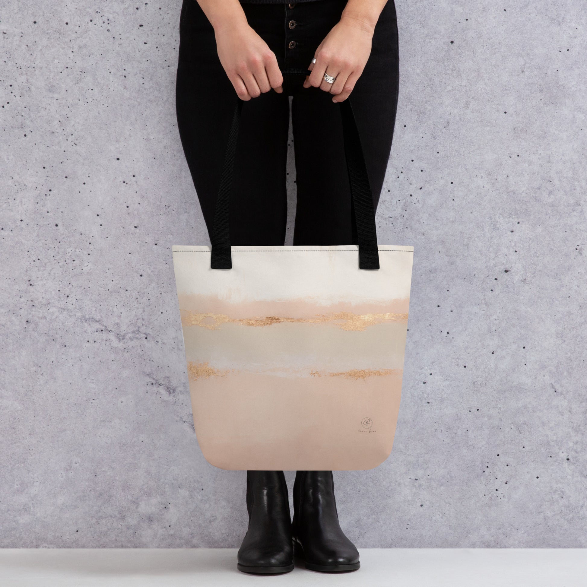 Person holding a beige and blush tote bag with gold brushstroke design and black straps, standing against a grey wall background.