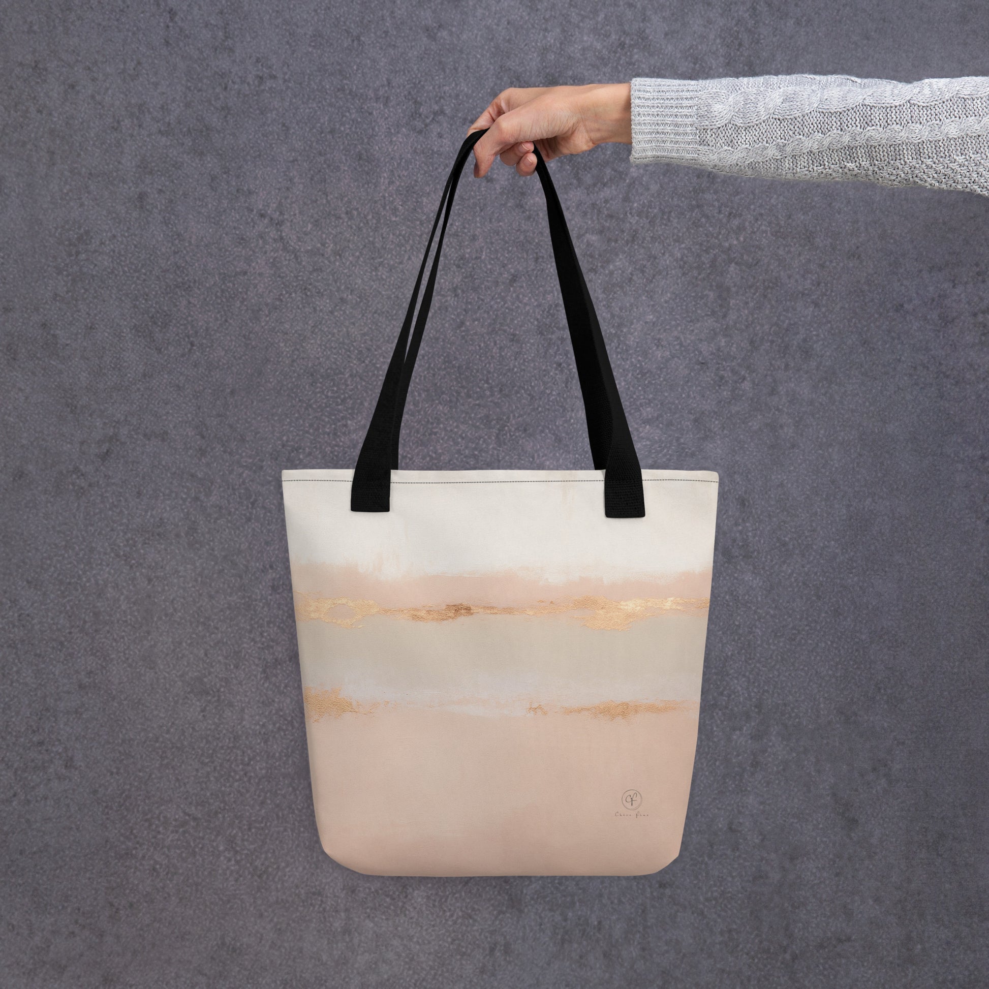 Hand holding a beige and blush tote bag with gold horizon-style brushstrokes and black straps, against a soft grey background.