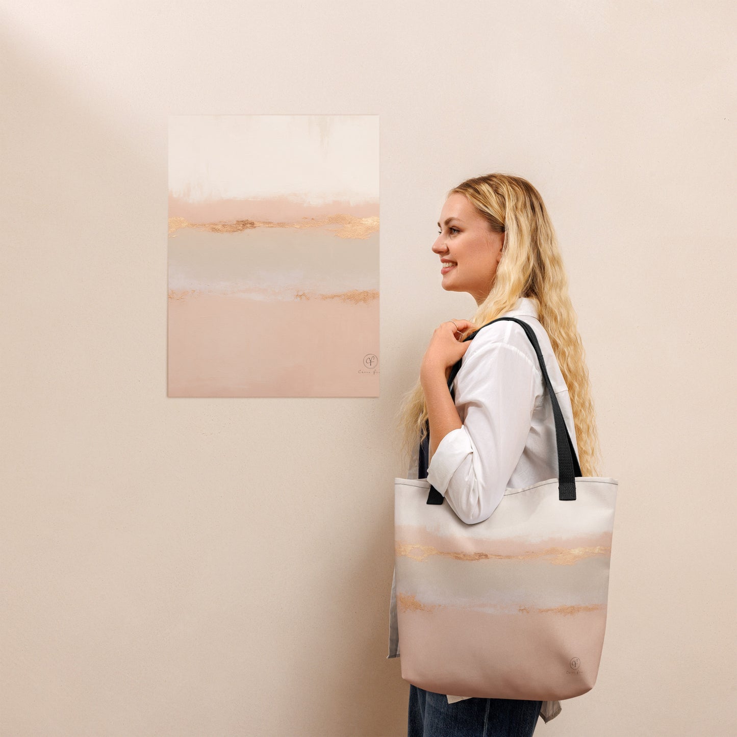 Smiling woman holding a beige and blush tote bag with gold horizon design, standing beside a matching abstract art print on a light beige wall.