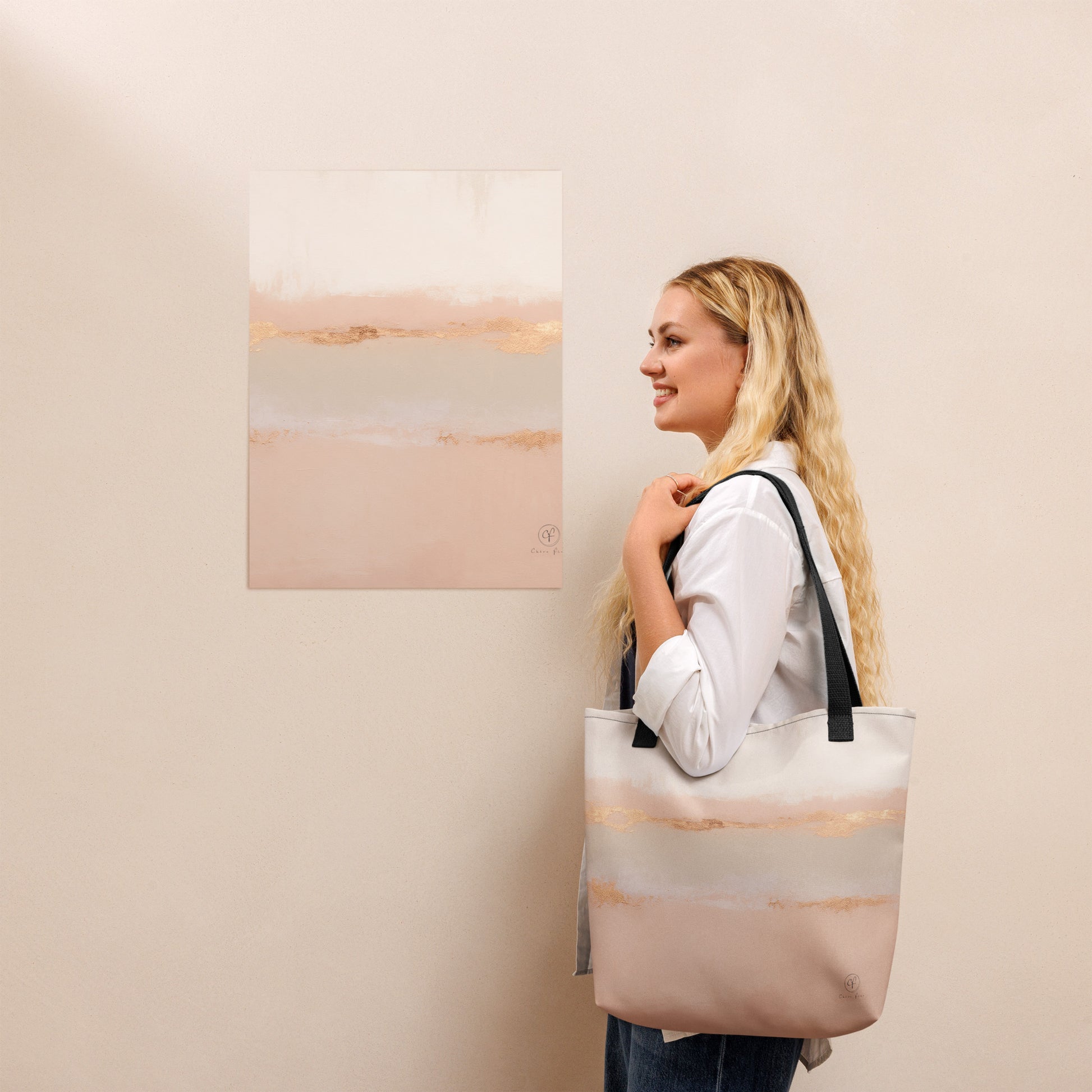 Smiling woman holding a beige and blush tote bag with gold horizon design, standing beside a matching abstract art print on a light beige wall.