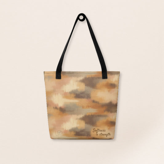 Beige and brown patterned tote bag with black handles and the phrase “Softness is strength” printed on the bottom right.