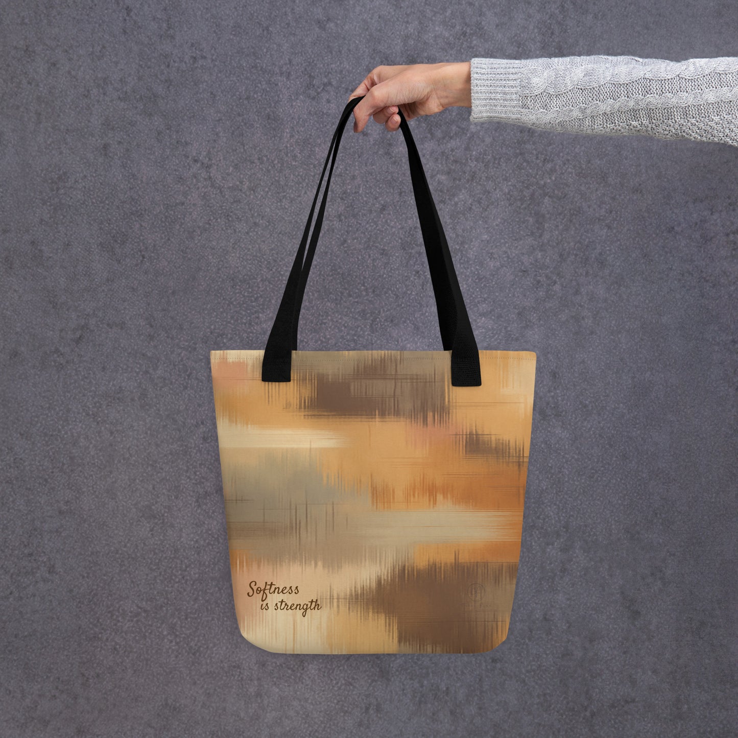 Hand holding a beige and brown patterned tote bag with black handles and the phrase “Softness is strength,” against a grey textured wall.