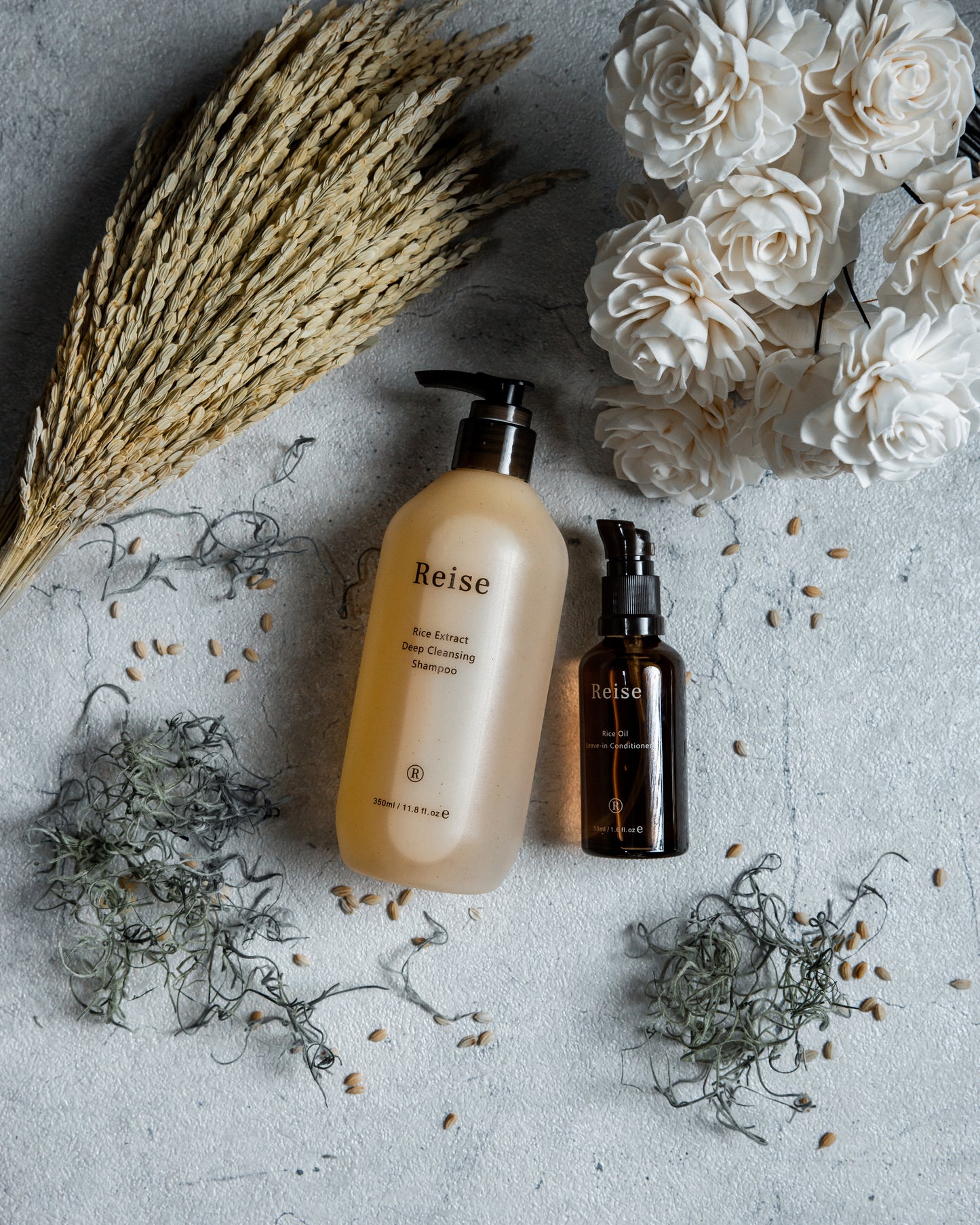Two bottles of Reise haircare products on a textured surface with flowers and herbs.